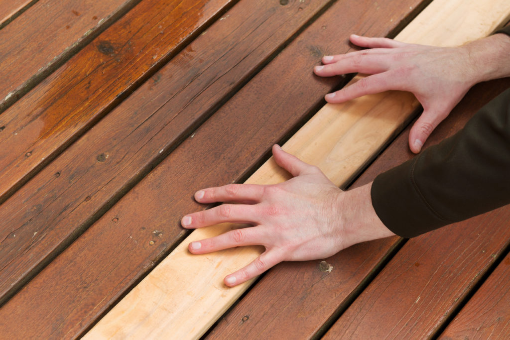 a brand new board on a deck lying amongst older boards with two hands on top holding it down is part of a carpentry repair